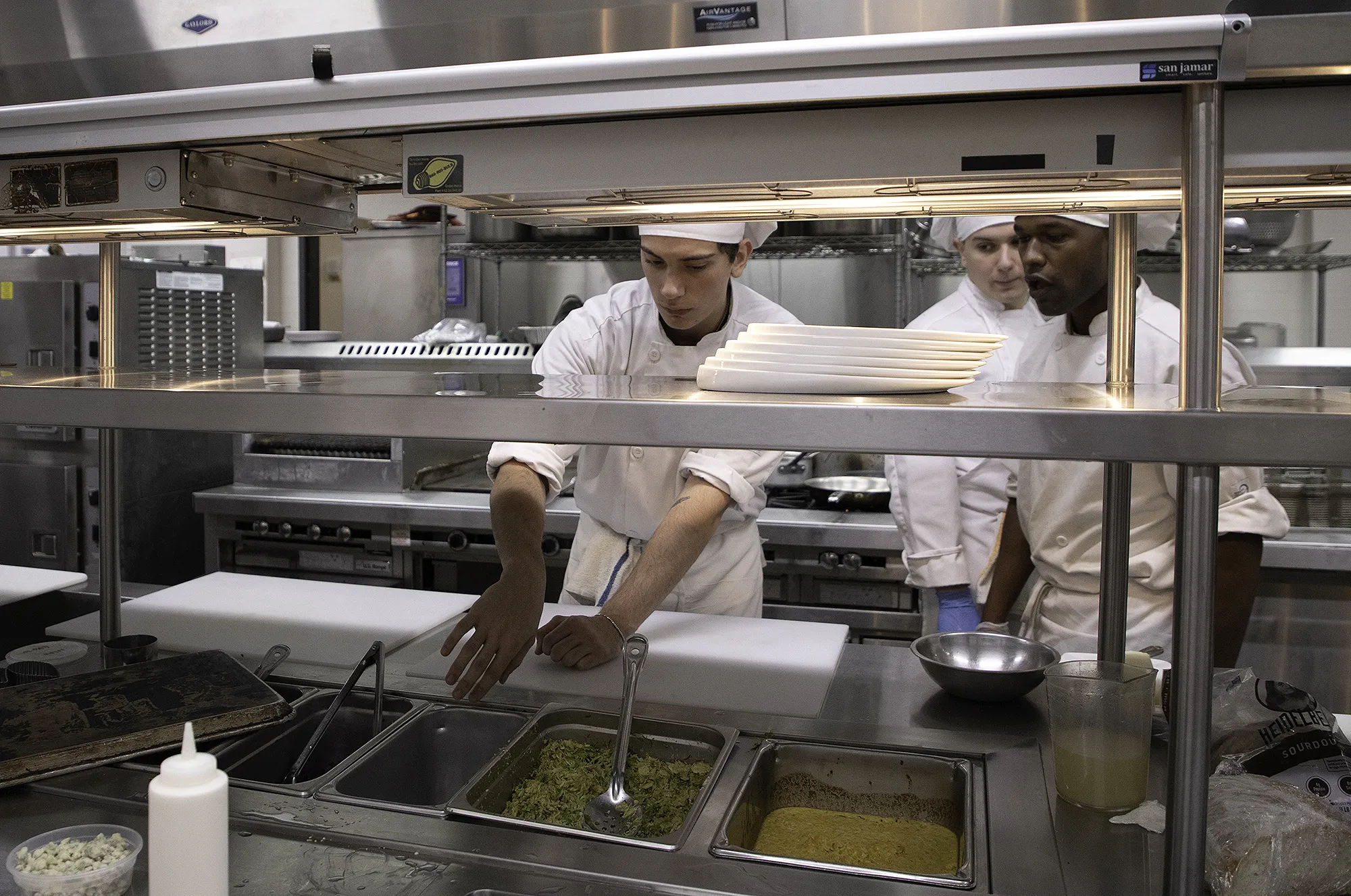 Chef Training Culinary arts students in white chef coats and hats prepare food at a stainless-steel service line, arranging ingredients and plates in a commercial teaching kitchen