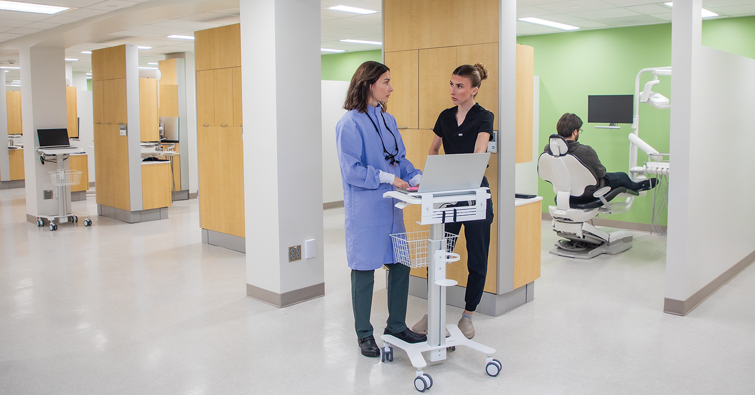 Two people standing by a workstation in a dental clinic with a patient sitting in a chair in the room behind them