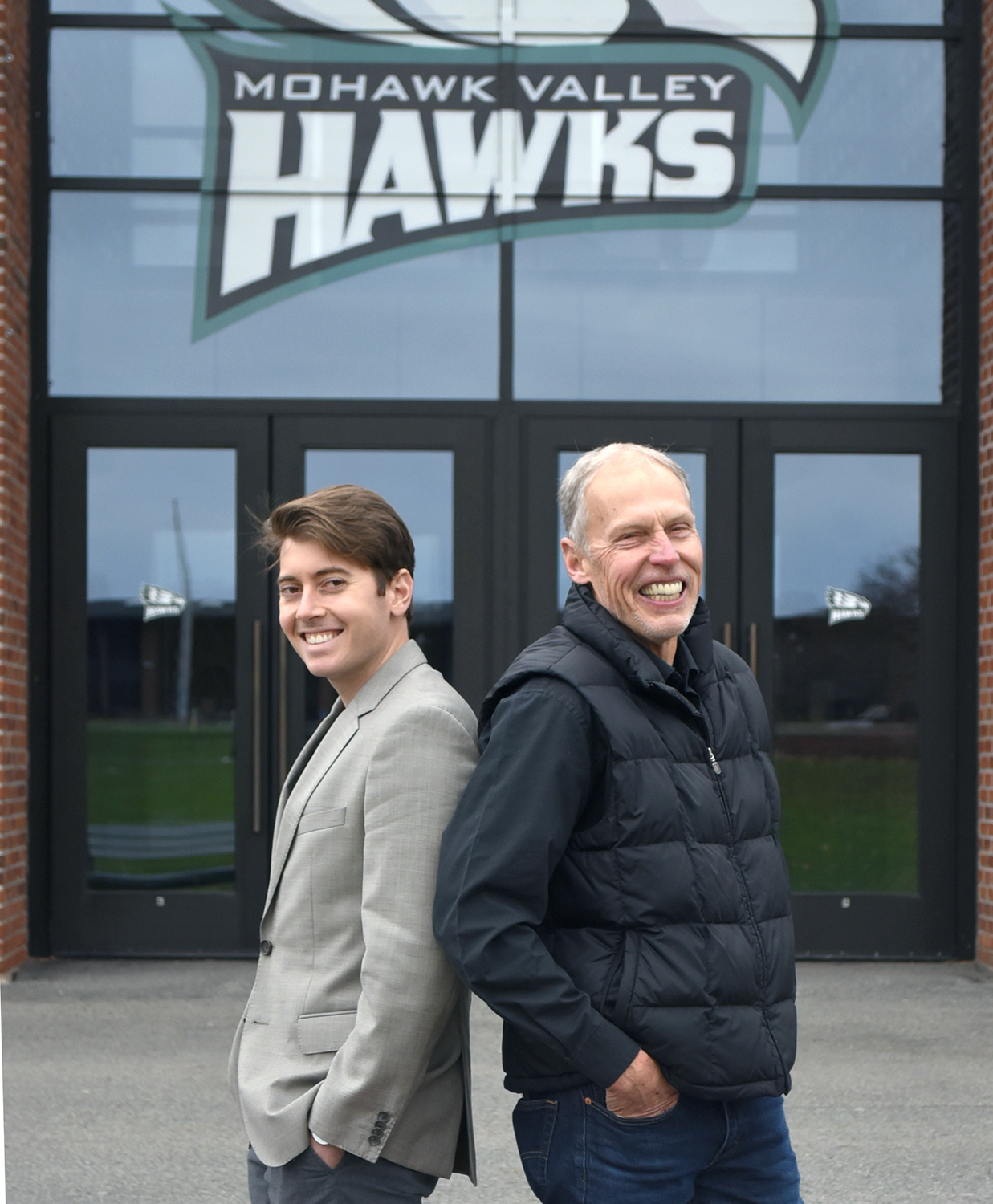 Image of David and Bob Gould standing back-to-back in front of the gym with Hawk logo