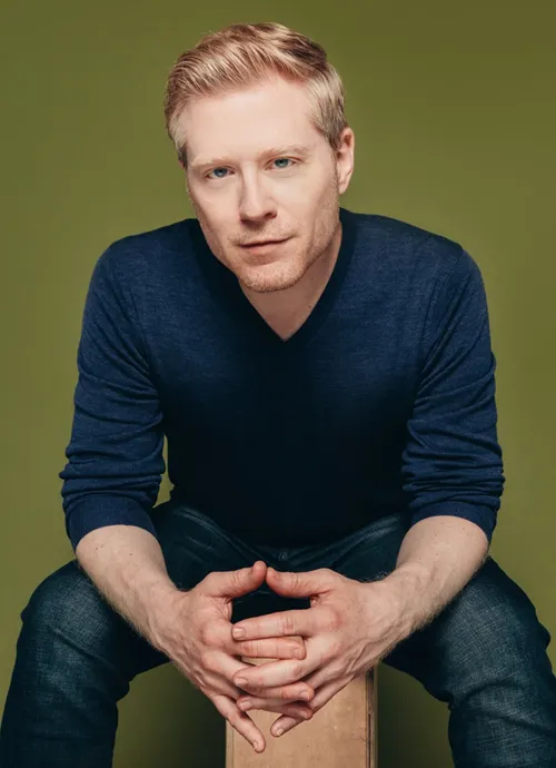 Image of Anthony Rapp sitting with hands together against green background