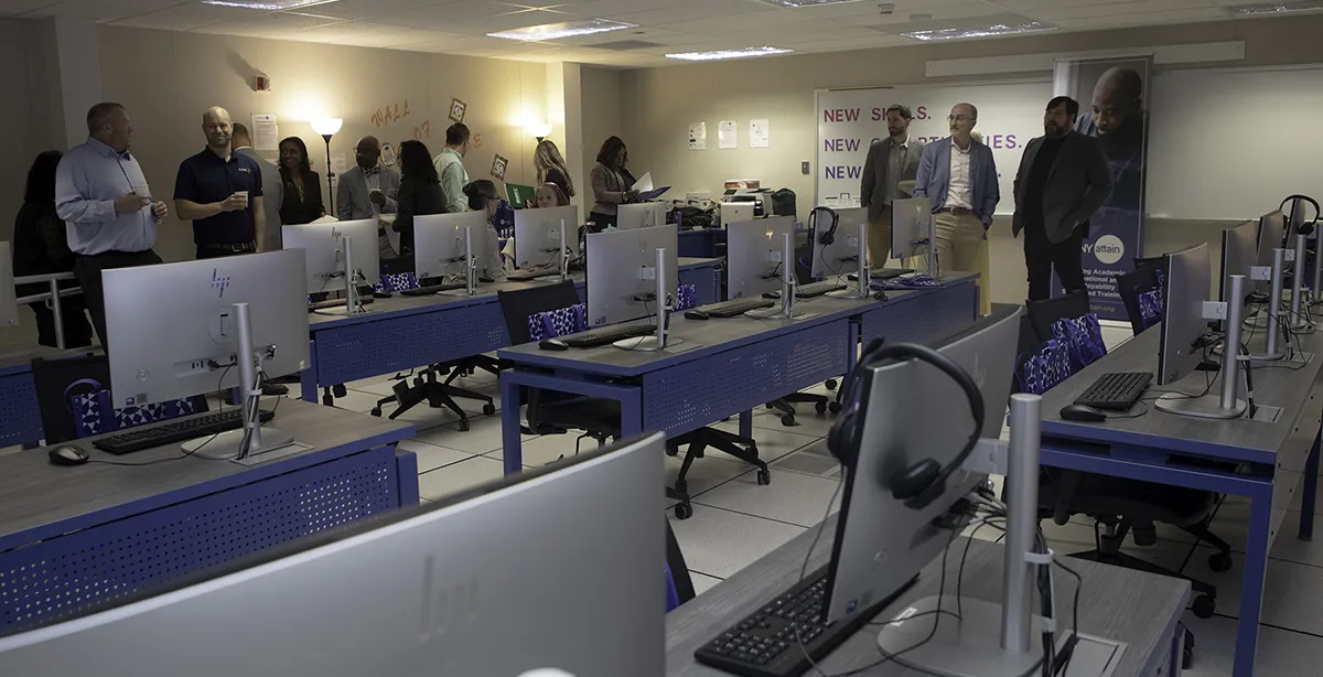 MVCC ATTAIN Lab Computer lab with blue desks and silver computers, with people standing in the background