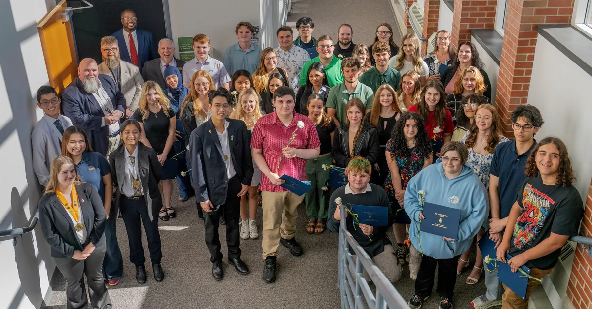 Large group of students and staff gathered on an indoor stairway, posing for a photo. Several students hold certificates and white flowers, and one student at the front wears a medal. The group is dressed in a mix of casual and formal clothing, smiling toward the camera in a bright hallway with brick walls and windows.