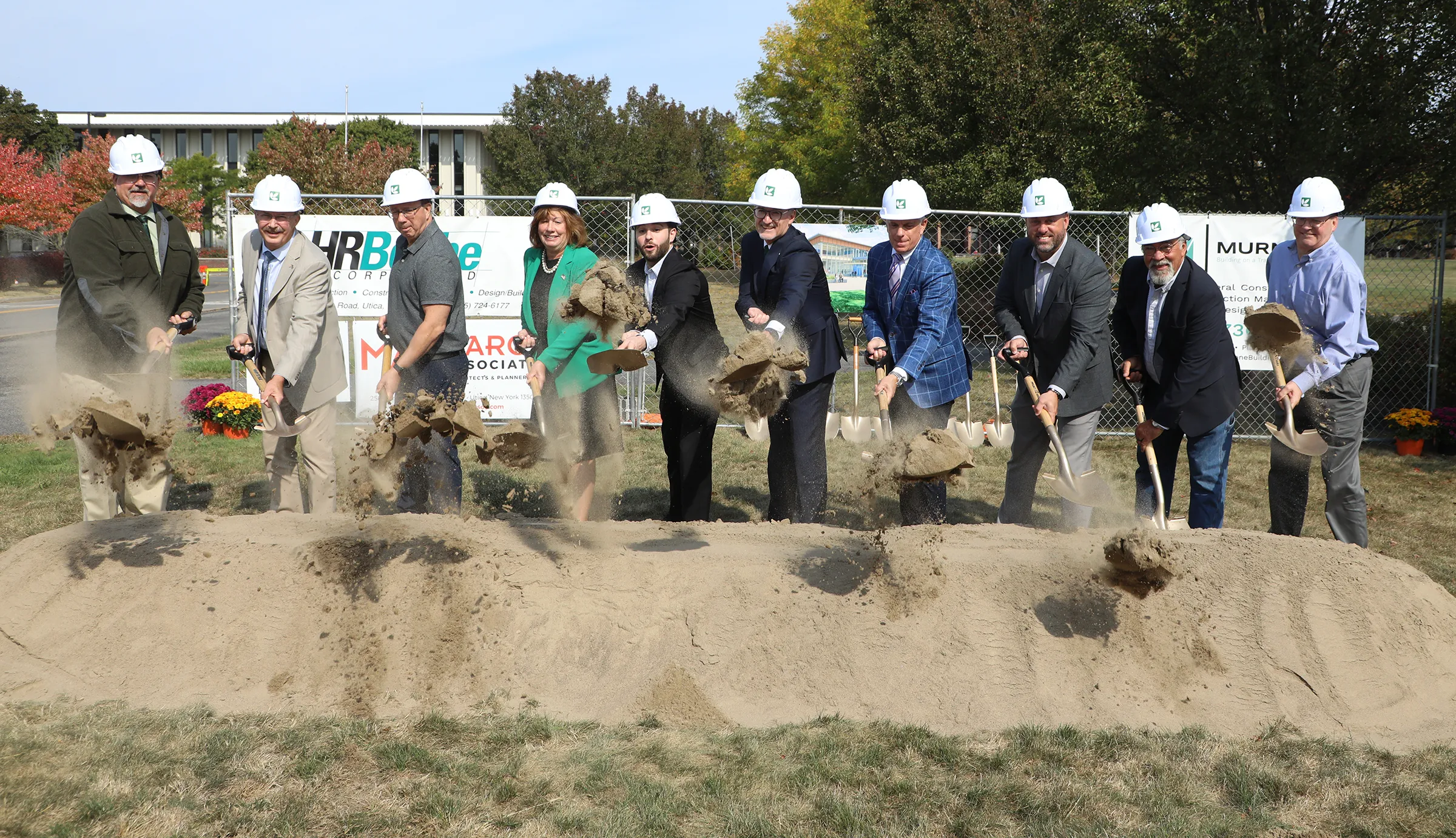 A group of people wearing white construction helmets use shovels to toss dirt during a groundbreaking ceremony outdoors. A banner with construction company logos is visible in the background.