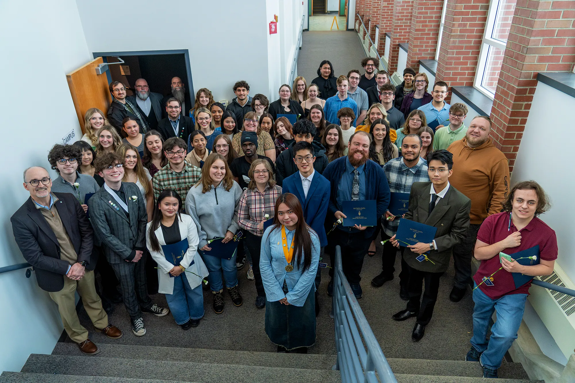 Spring 2026 PTK Large group of students and staff gathered on an indoor stairway, posing for a photo. Several students hold certificates and white flowers, and one student at the front wears a medal. The group is dressed in a mix of casual and formal clothing, smiling toward the camera in a bright hallway with brick walls and windows.